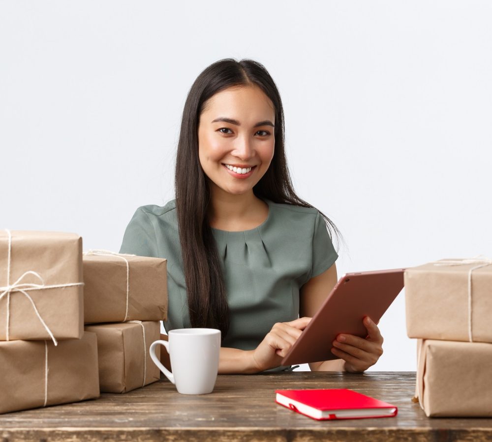 Small business owners, startup and e-commerce concept. Smiling asian businesswoman work from home, packing sold items, wrap boxes with orders for shipping, manage online store via digital tablet.
